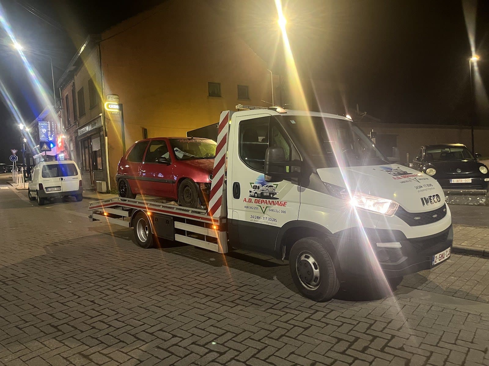 Le camion plateau Iveco d’A.D. Dépannage chargé d’une Citroën, de nuit, phares allumés